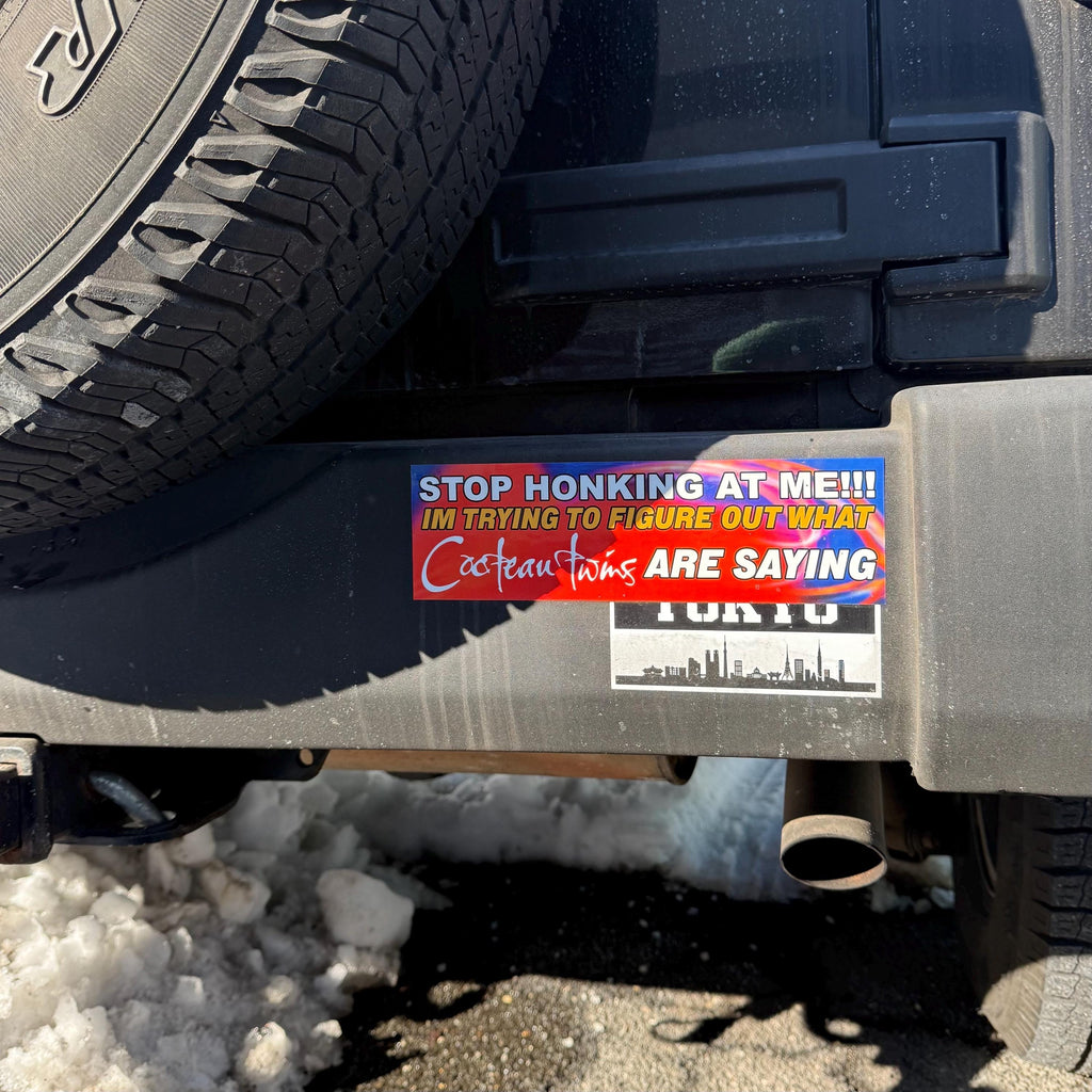 Bumper sticker on a car reading 'Stop honking at me! I'm trying to figure out what Cocteau twins are saying' with a snowy background.
