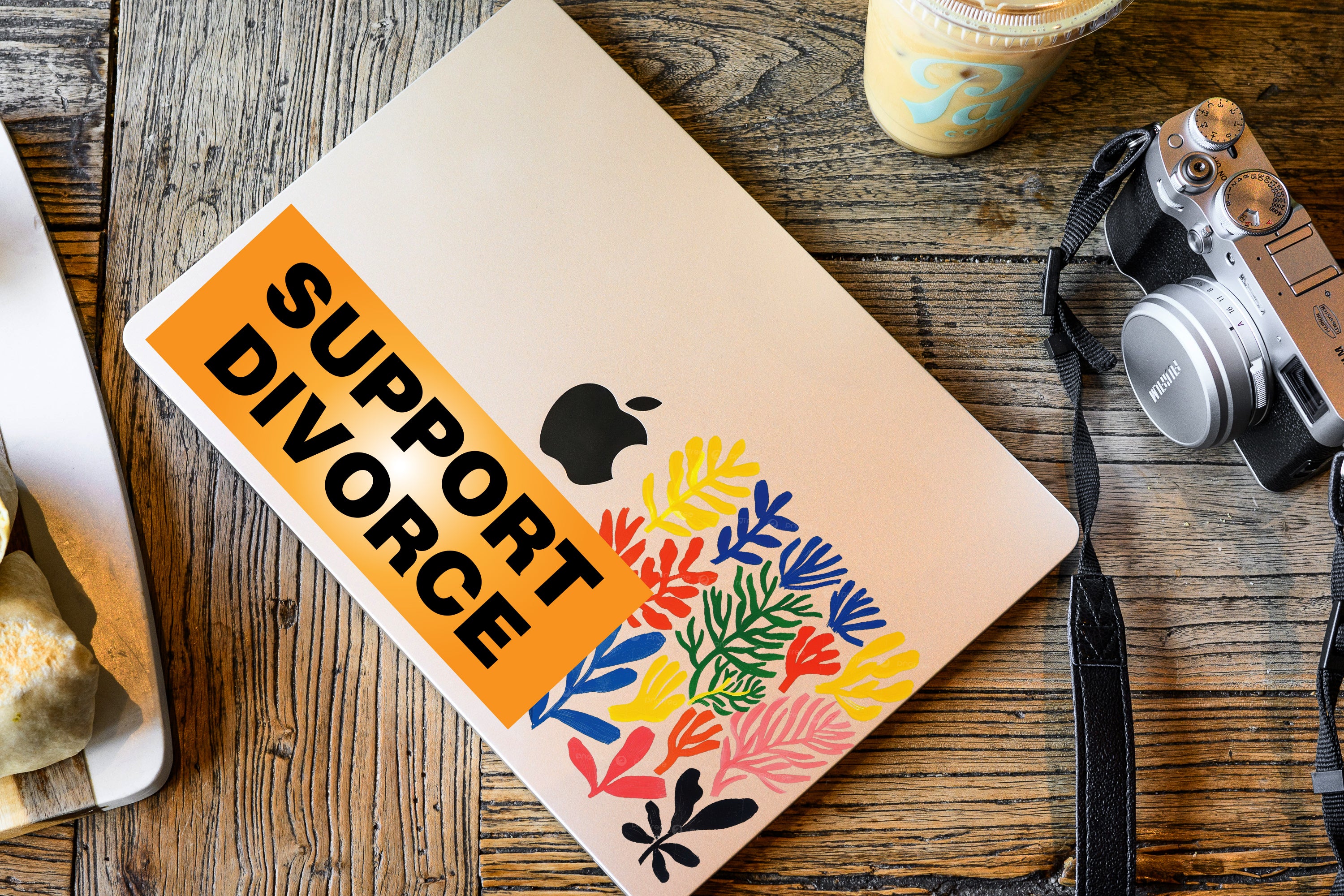 Notebook with 'Support Divorce' sticker on a wooden surface with a camera and coffee cup.